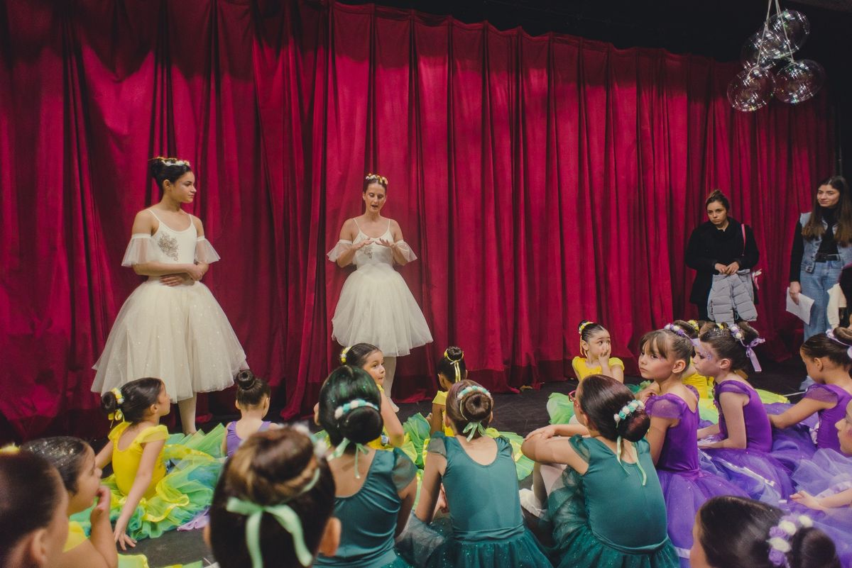 Bailarinas de tutu branco diante da cortina vermelha, com as turmas infantis sentadas à frente