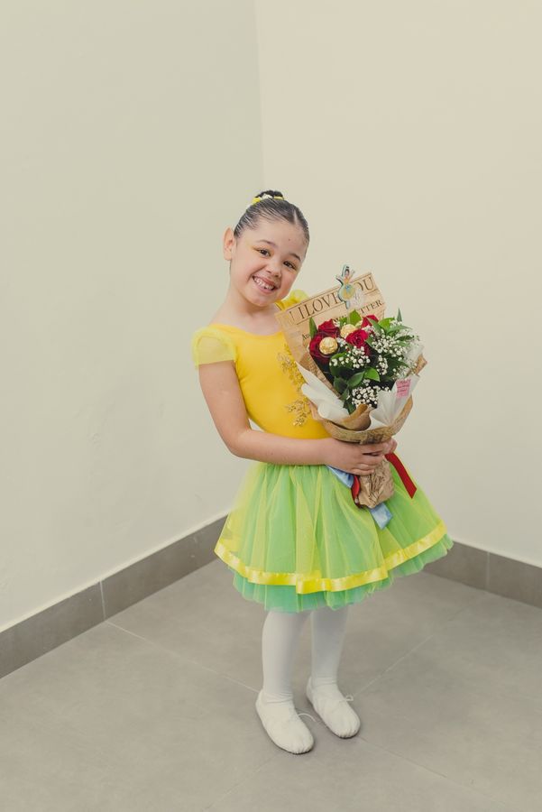 Aluna pequena de collant amarelo e saia verde sorrindo com um buquê de flores