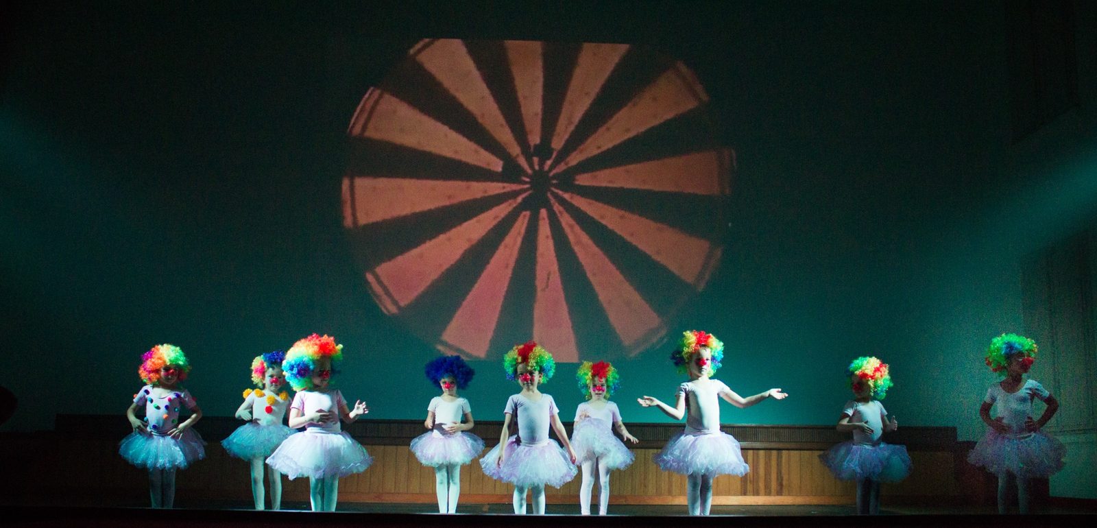 Turma infantil da Brilhart em cena no palco, de tutu e perucas coloridas