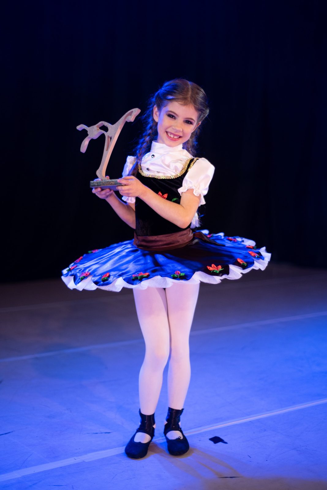 Aluna da Brilhart sorrindo no palco, de figurino azul, segurando um troféu