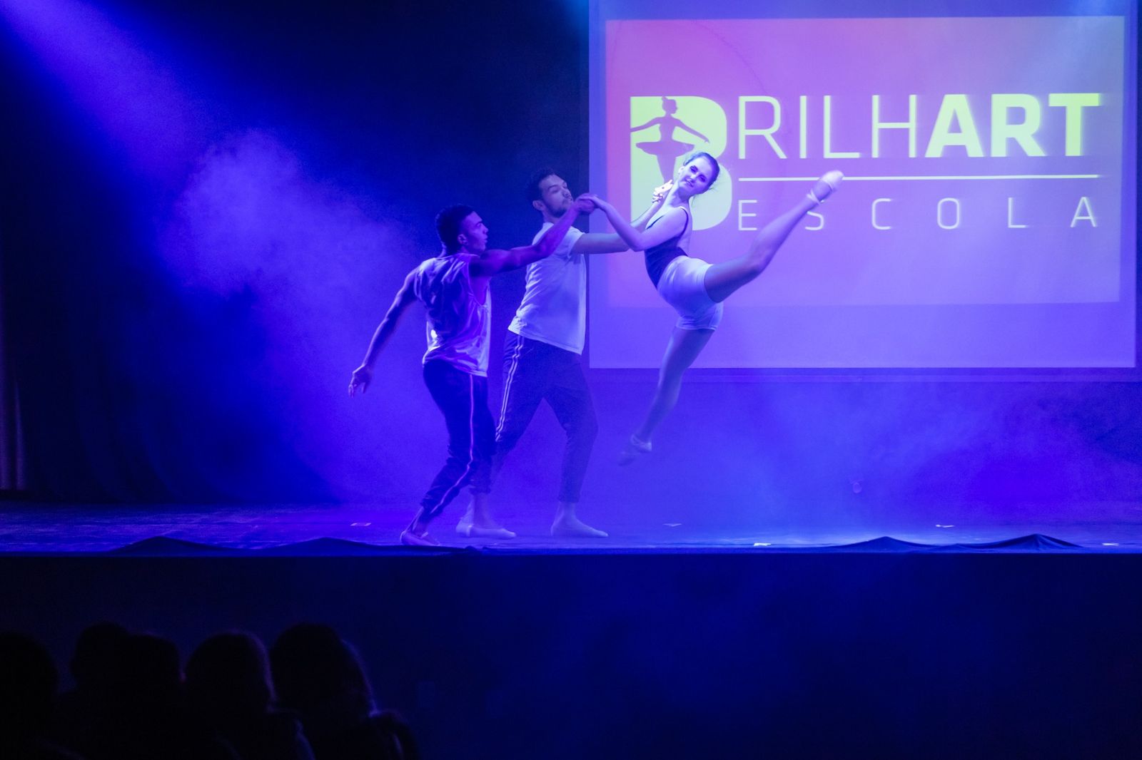 Três bailarinos da Brilhart em cena no palco, sob luz roxa, com o logo da escola projetado ao fundo