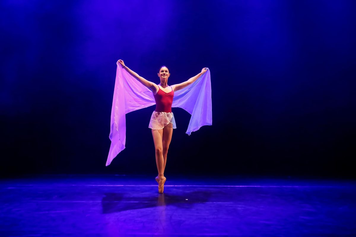 Larissa Cléria no palco, de collant vermelho, abrindo um tecido lilás com os braços